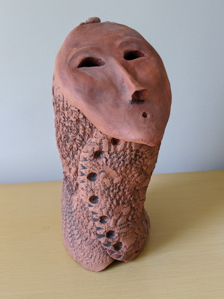 orange clay sculpture of serene oval face with a curved nose, half moon eyes, and a small mouth singing singing in an O
