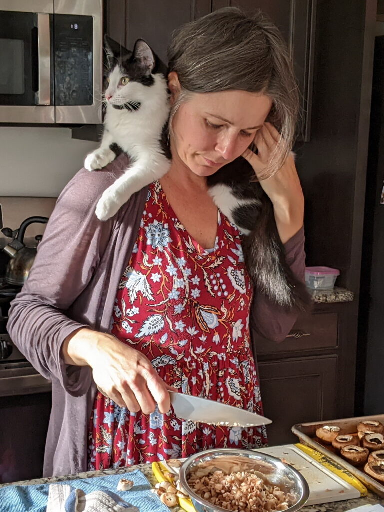 white woman in red dress with blue flowers in the kitchen chopping mushrooms with black and white cat on her shoulders staring out the window