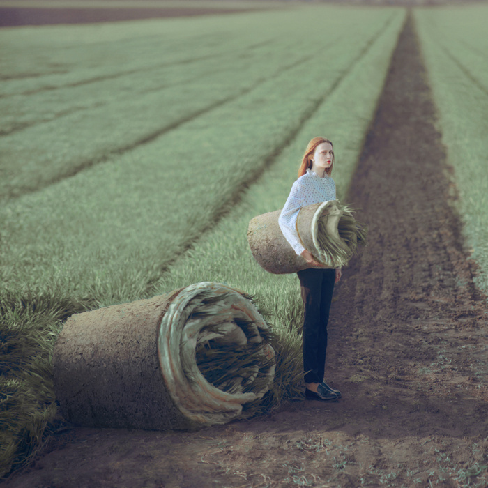 woman with red hair and a stern look on her face stands in a wheat field holding a coiled row of wheat like it's a blanket