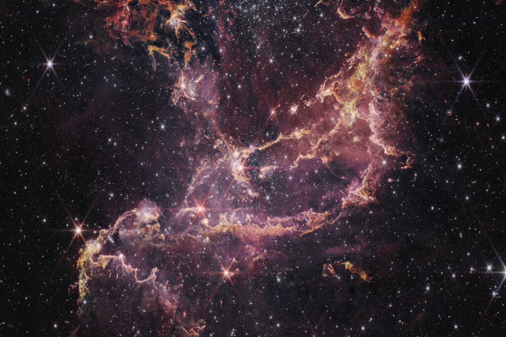 A star cluster within a nebula. The center of the image contains arcs of orange and pink gas that form a boat-like shape. One end of these arcs points to the top right of the image, while the other end points toward the bottom left. Another plume of orange and pink gas expands from the center to the top left of the image. To the right of this plume is a large cluster of white stars. There are more of these white stars and galaxies of different sizes spread throughout the image.