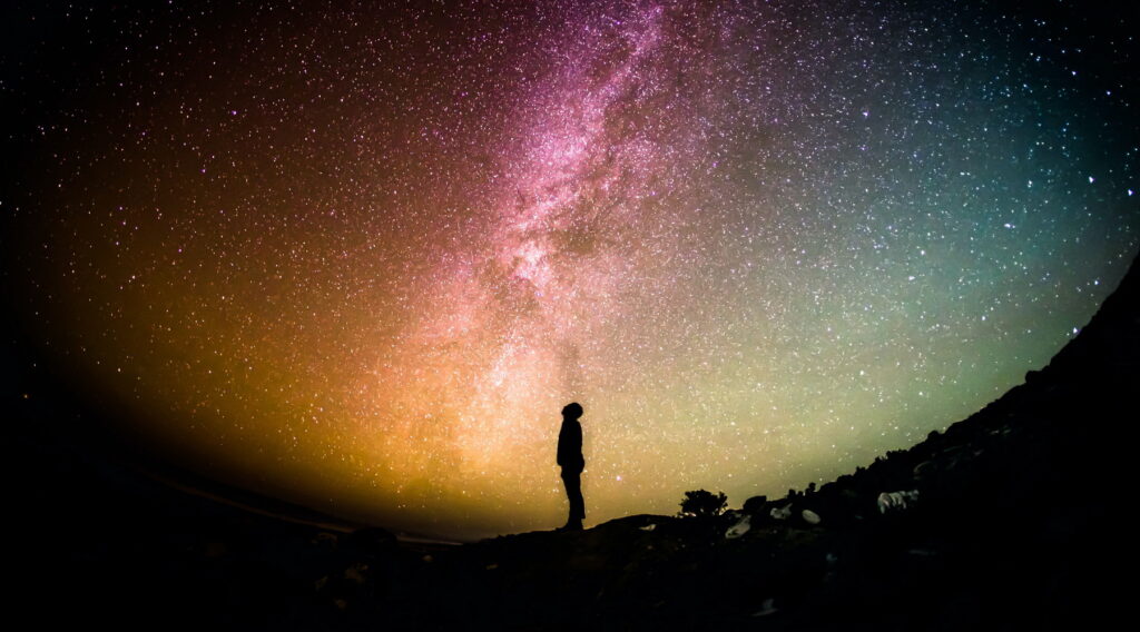 digital art of a silhouette of a person standing and looking up with a background of red, orange, green, blue blending together with what looks like start and a milky way
