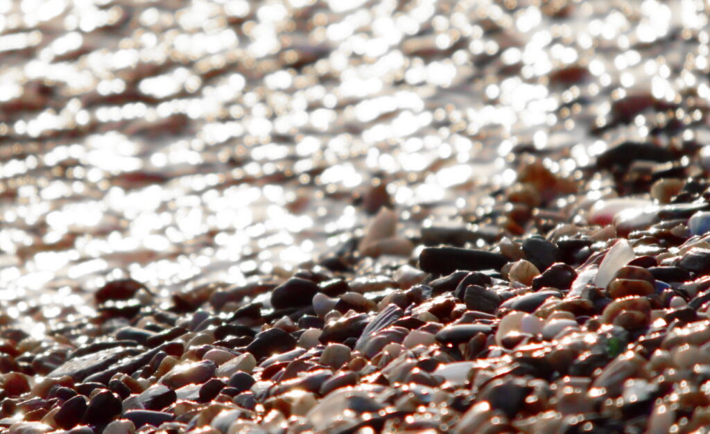 small colorful stones on the bank of rippling water topped with a glimmer of sunlight