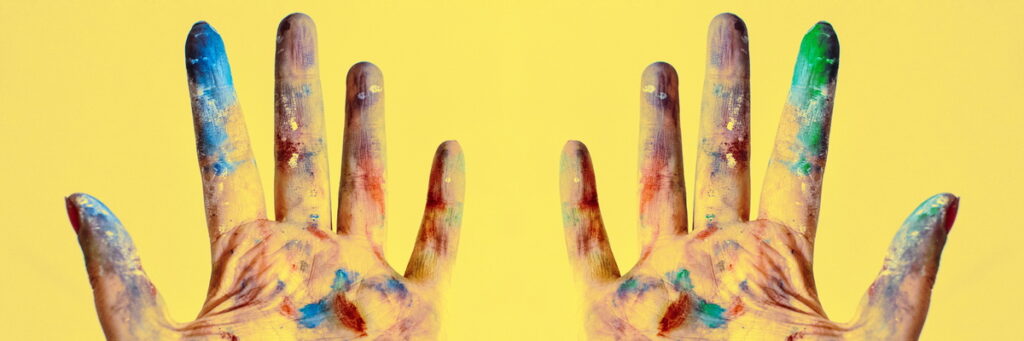 two hands dirty with multi-colored paint, palms up, fingers spread wide, over bright yellow background