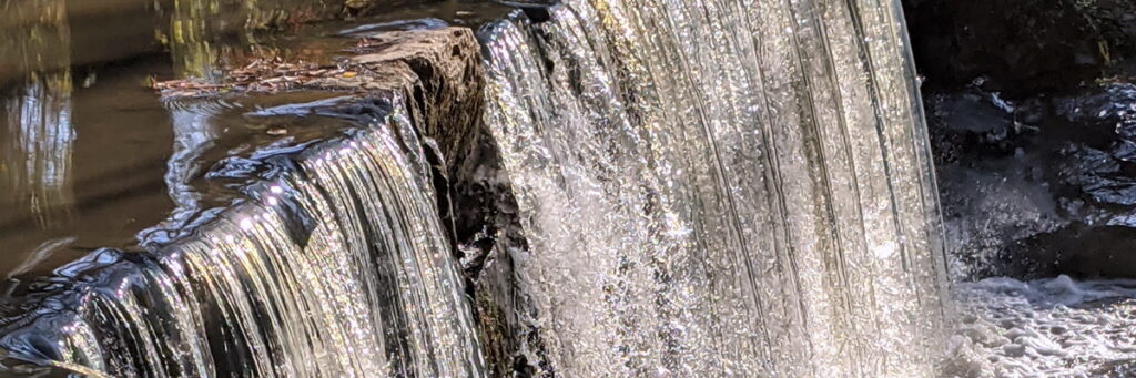 close up: the edge of a small spillway with calm water on top dropping over in a curtain of water sparkling in the sun