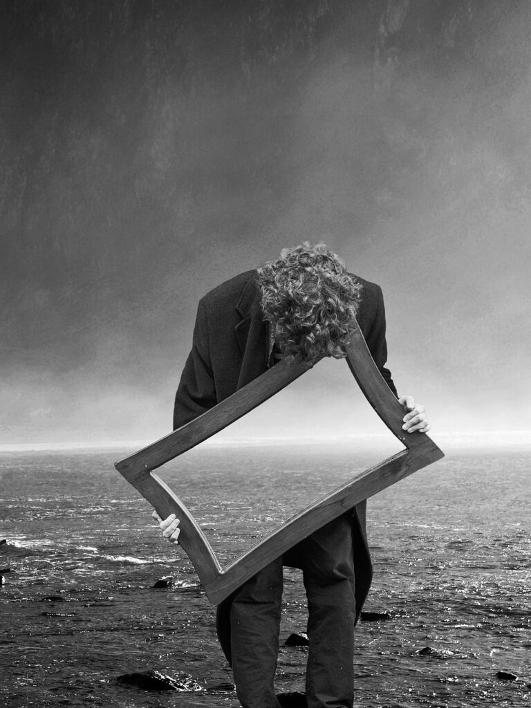 Black and white abstract photo of a person with mid-length curly hair, wearing a suit, standing on the edge of choppy waters, bent over with forehead on a rectangle-shaped mirror reflecting the water behind him