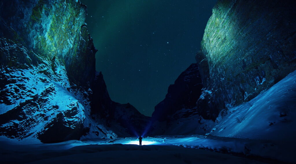Two mountain walls with a starry night sky between them, and a person on the ground with lights shining on the mountains reflecting greens and blues, the ground covered in white snow